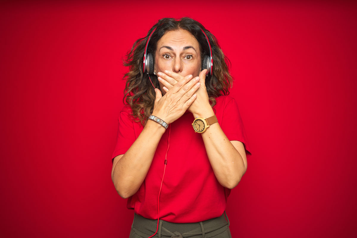 women with headphones on, in front of red background, with hands over her mouth as if she said the wrong thing